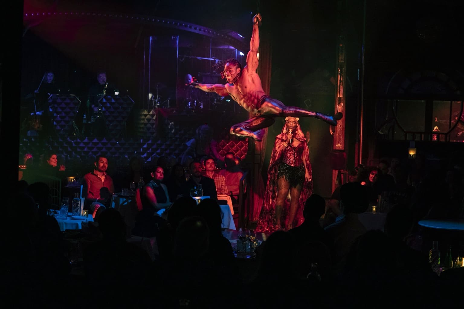 Alseny suspended one-armed in a mid-air split above a seated theatre audience during a live stage performance, with a vocalist and band on stage