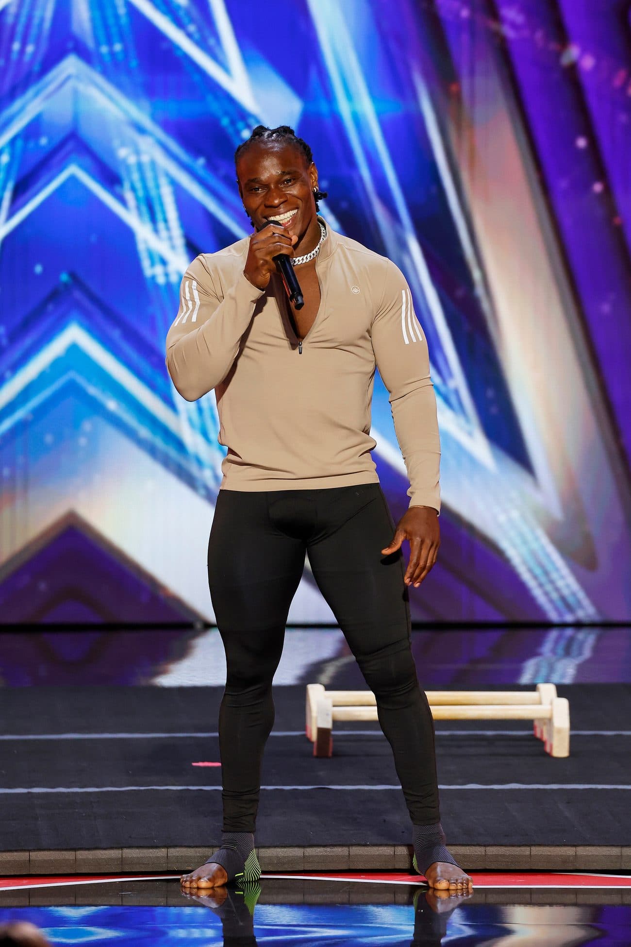 Alseny Sylla on the America's Got Talent stage holding a microphone, with parallettes on the floor