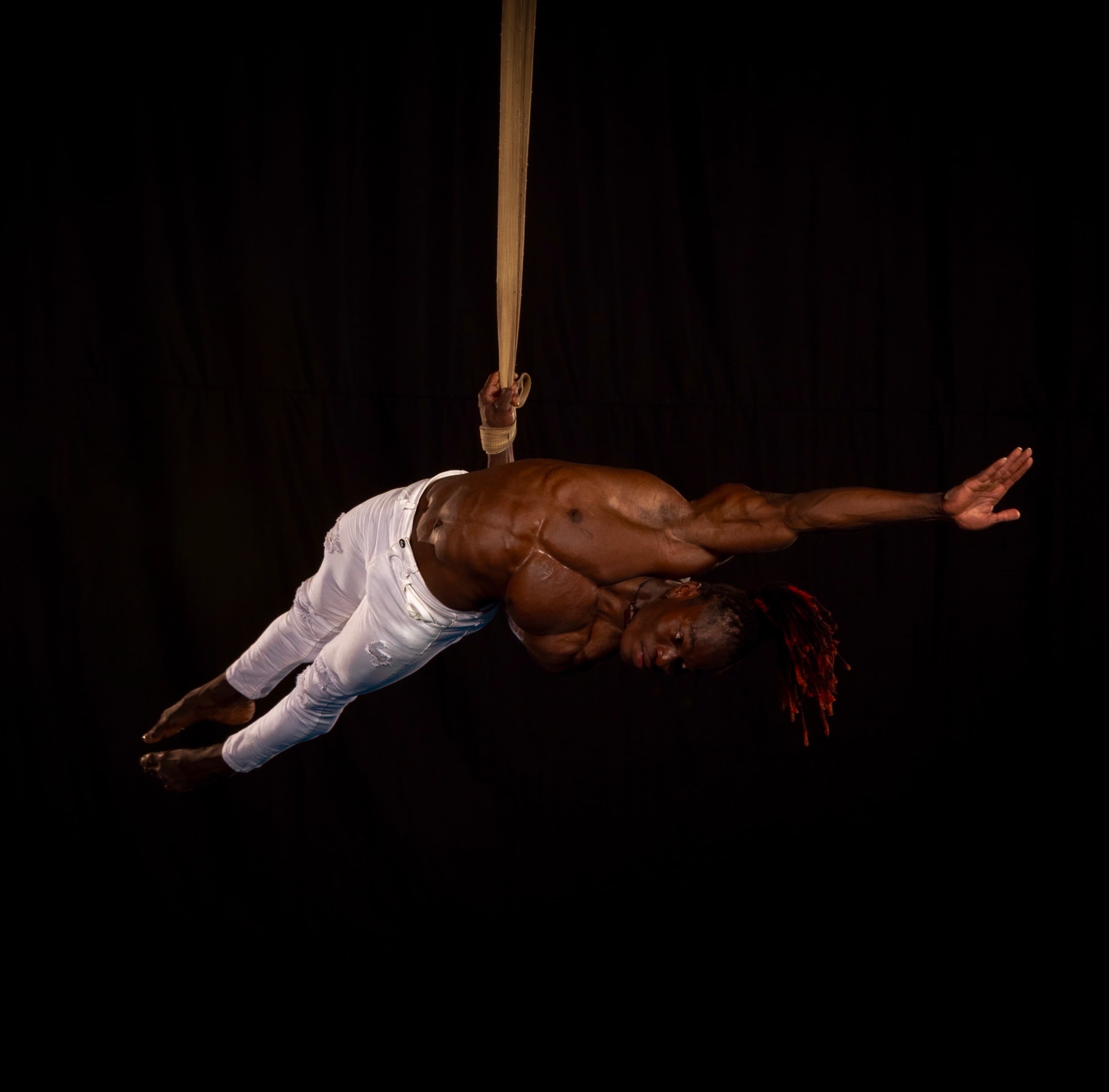 Alseny Sylla holding a horizontal one-arm hold on a single aerial strap against a black backdrop